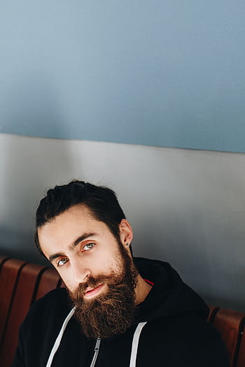 Close-up portrait of a bearded man with green eyes posing indoors against a blue wall. HD phone wallpaper