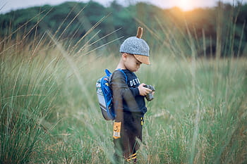 A young boy wearing a cap explores a grassy field with a camera, enjoying outdoor adventure. HD wallpaper