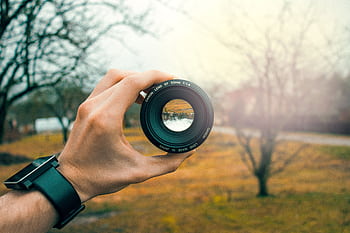 Close-up of a hand holding a 50mm camera lens outdoors, focusing through the lens. HD wallpaper