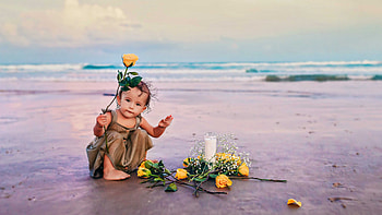 Cute Toddler Girl Baby With Yellow Flowers Is Sitting On Beach Sand Wearing Brown Color Dress In Blur Ocean Waves Background Cute HD wallpaper