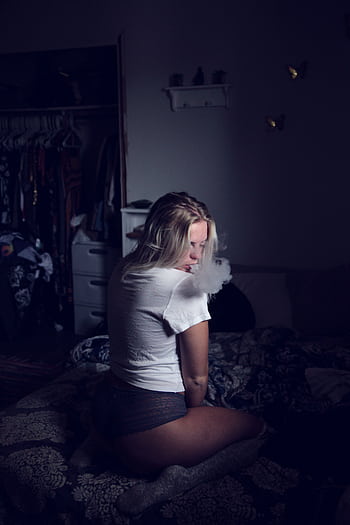 A young woman with blond hair smoking in a dimly lit bedroom, creating a moody atmosphere. HD phone wallpaper