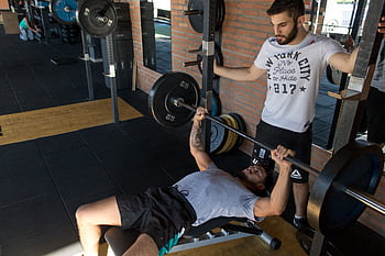 A strong man lifts weights on a bench while a trainer assists in a modern gym setup. HD wallpaper