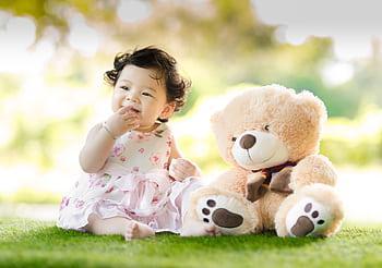 Smiling baby girl sits on grass with a plush teddy bear, enjoying a sunny day. HD wallpaper