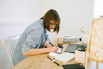 A young woman working from home, writing notes at a desk with a laptop and papers. HD wallpaper