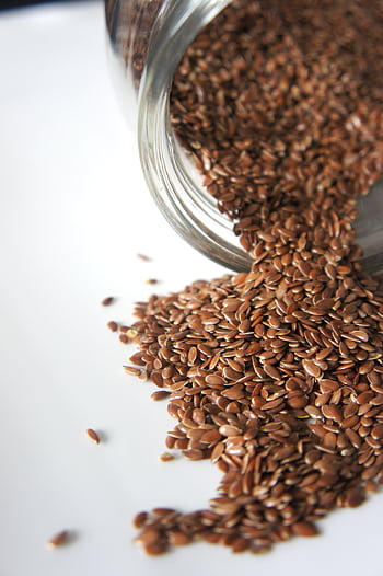 High angle view of flaxseeds spilling from a jar onto a white surface. Perfect for health and food concepts. HD phone wallpaper