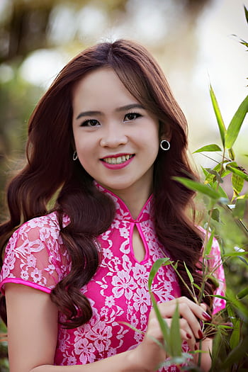 A smiling woman in a pink floral dress poses outdoors surrounded by greenery. HD phone wallpaper