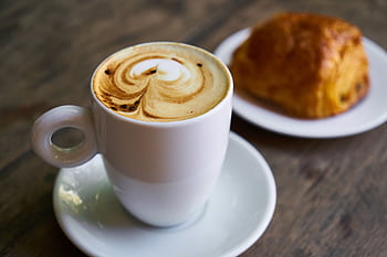 A delicious cappuccino with latte art next to a pastry on a wooden table, perfect for breakfast moments. HD wallpaper