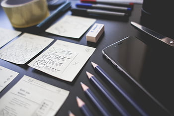A creative workspace featuring UI design plans, pencils, a smart, and other office tools. HD wallpaper