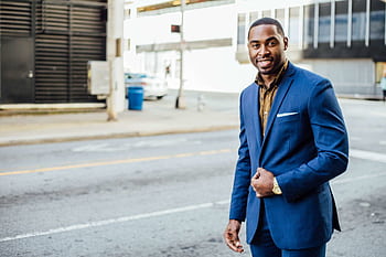 A confident businessman in a blue suit smiling outdoors in an urban setting during the day. HD wallpaper