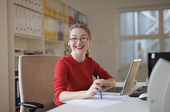 Happy woman in red sweater working at office desk with laptop and pen, smiling at camera. HD wallpaper