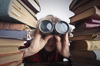 A man observing through binoculars surrounded by stacks of vintage books indoors. HD wallpaper