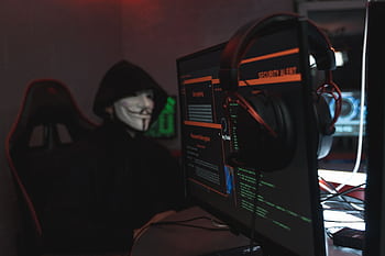 A person wearing a Guy Fawkes mask engaged in hacking activities on a computer in a dimly lit room. HD wallpaper