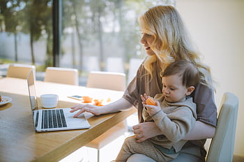 A mother works on a laptop at home while holding her baby, showcasing remote working and parenting. HD wallpaper