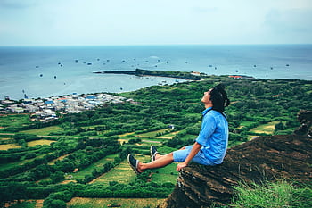 A young man sits peacefully on a cliff, enjoying the scenic ocean view. HD wallpaper