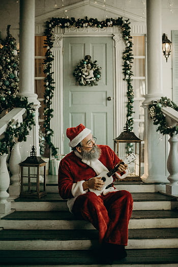 Santa Claus sitting on decorated porch steps, playing ukulele, surrounded by Christmas lights and greenery. HD phone wallpaper