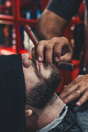 A barber skillfully shaves a man's beard using a straight razor at a barbershop. HD phone wallpaper