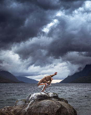 Person posing energetically on a rock by the water with dramatic storm clouds above, creating a powerful and dynamic scene. HD phone wallpaper