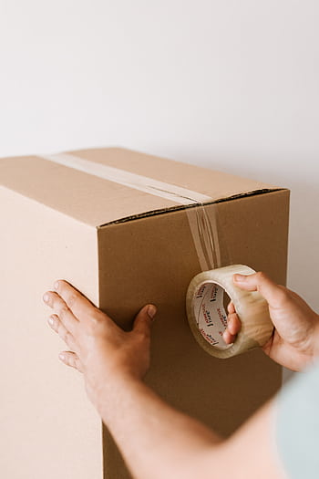 Close-up of hands using tape to seal a cardboard box, ideal for moving or shipping themes. HD phone wallpaper