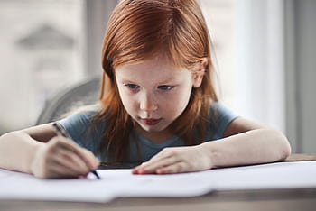 Focused young girl with red hair writing on paper at a desk indoors. HD wallpaper