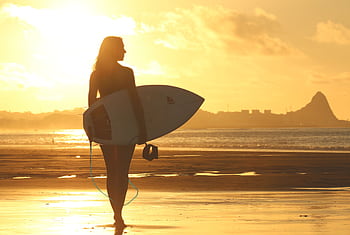 A silhouette of a woman holding a surfboard, standing on a beach at sunset with a golden sky. HD wallpaper