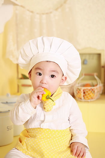 Cute baby in chef hat and apron nibbling a bell pepper in bright kitchen. HD phone wallpaper