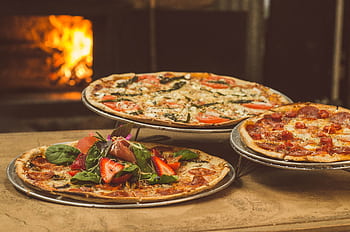 Trio of gourmet pizzas with unique toppings near a wood-fired oven, ideal for food enthusiasts. HD wallpaper