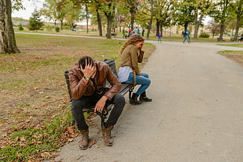 Distraught couple sitting apart on a park bench, facing away from each other, in a public park setting. HD wallpaper