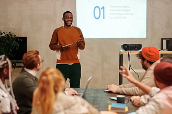 A man delivers a presentation in a modern office to a diverse group of colleagues. HD wallpaper