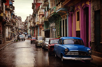 Colorful vintage cars line a wet street with vibrant buildings in a cityscape setting. HD wallpaper