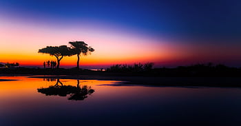 Vibrant sunset with silhouettes reflected in calm water creating a serene landscape. HD wallpaper