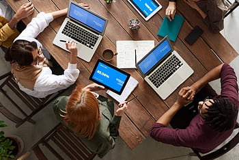 Diverse team discussing business strategies with laptops and tablets at a wooden table. HD wallpaper