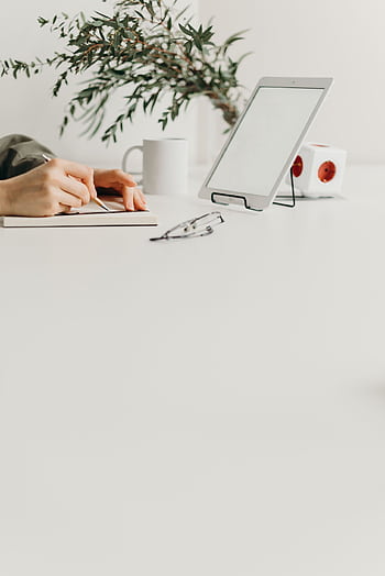 A clean, minimalist workspace with a tablet, plant, and hand writing in a notebook. HD phone wallpaper