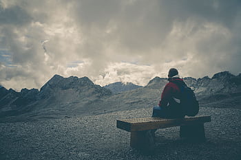 A solitary traveler sits on a bench, gazing at a mountain range under a cloudy sky. HD wallpaper