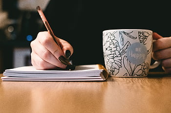 Person writing in a notebook with a floral ceramic mug on a wooden desk. HD wallpaper