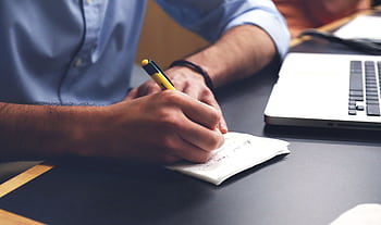 Close-up of a man writing notes with a pen next to a laptop on a desk. HD wallpaper