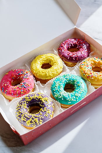 Brightly colored glazed donuts with sprinkles in a box, captured on a marble background, showcasing their vibrant colors. HD phone wallpaper