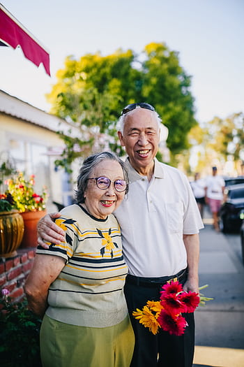 A happy elderly couple embracing outdoors, holding flowers on a sunny day. HD phone wallpaper