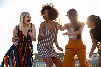 Four diverse women enjoying drinks and laughter on a sunny rooftop. HD wallpaper