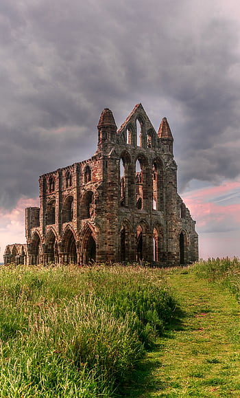 Dramatic view of the ancient Gothic ruins of Whitby Abbey under a cloudy sunset sky, surrounded by lush grass. HD phone wallpaper