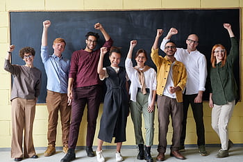 A group of eight adults cheerfully standing with raised fists in front of a blackboard indoors. HD wallpaper