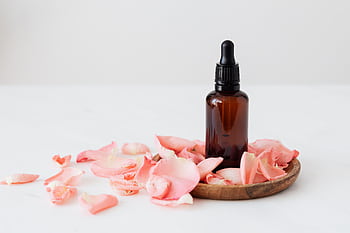 A brown dropper bottle surrounded by pink rose petals arranged on a wooden plate against a soft background. HD wallpaper