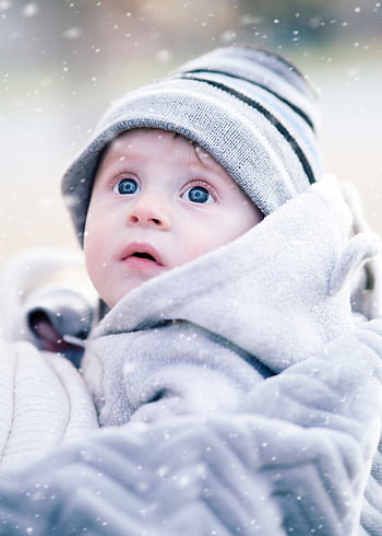 Cute baby wrapped in a blanket with a knit hat, gazing upward during snowfall. HD phone wallpaper