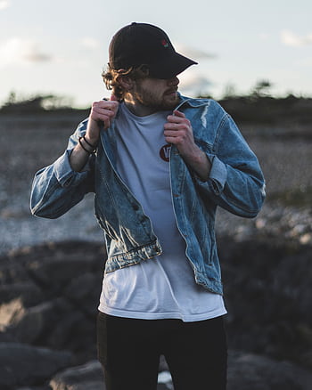 Man in a denim jacket and cap on a rocky outdoors setting, showcasing casual fashion. HD phone wallpaper