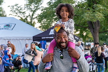 A joyful father carrying his daughter at an outdoor festival surrounded by people and tents. HD wallpaper