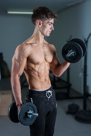 Muscular young man lifting dumbbells in a gym, showcasing strength and fitness. HD phone wallpaper