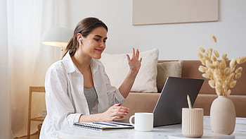 Cheerful young woman using laptop for a video call in a cozy living room, feeling connected and happy. HD wallpaper