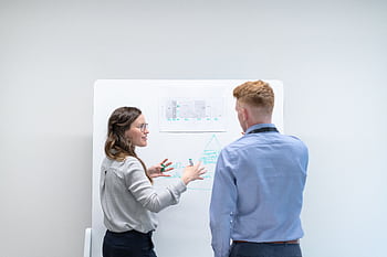 Two professionals discussing project plans at whiteboard in office setting. HD wallpaper