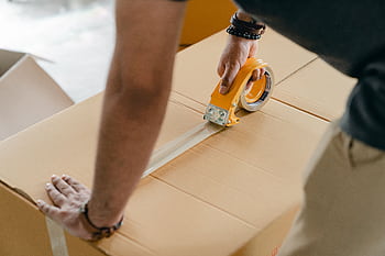 Man using packing tape to seal a cardboard box indoors. Ideal for moving or shipping concepts. HD wallpaper