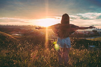 Woman with open arms enjoying sunset view over cityscape from a grassy hilltop. HD wallpaper