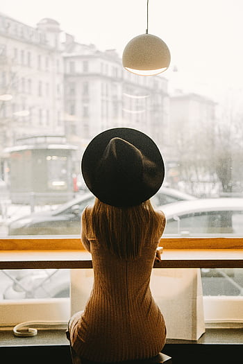 Stylish woman in a hat sits in a café, gazing outside at a bustling St. Petersburg street. HD phone wallpaper